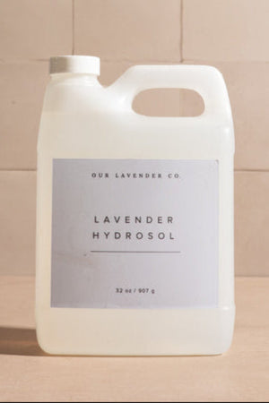 A large translucent white plastic jug of Our Lavender Co. Lavender Hydrosol with a lavender colored label, set against a background of beige square tiles.